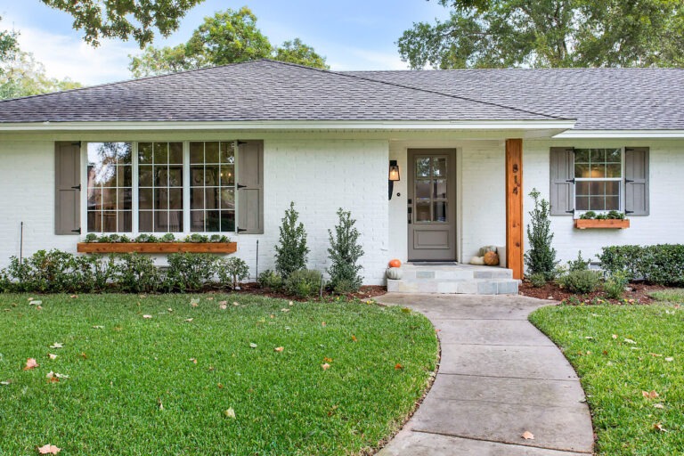 Charming white brick house with gray shutters, a wooden accent by the door, and landscaped front yard featuring shrubs and seasonal decor.