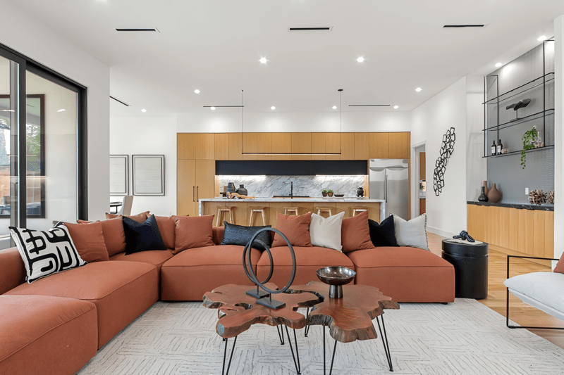 Spacious modern living room featuring a large orange sectional sofa, a unique coffee table, and an open kitchen with wooden accents.
