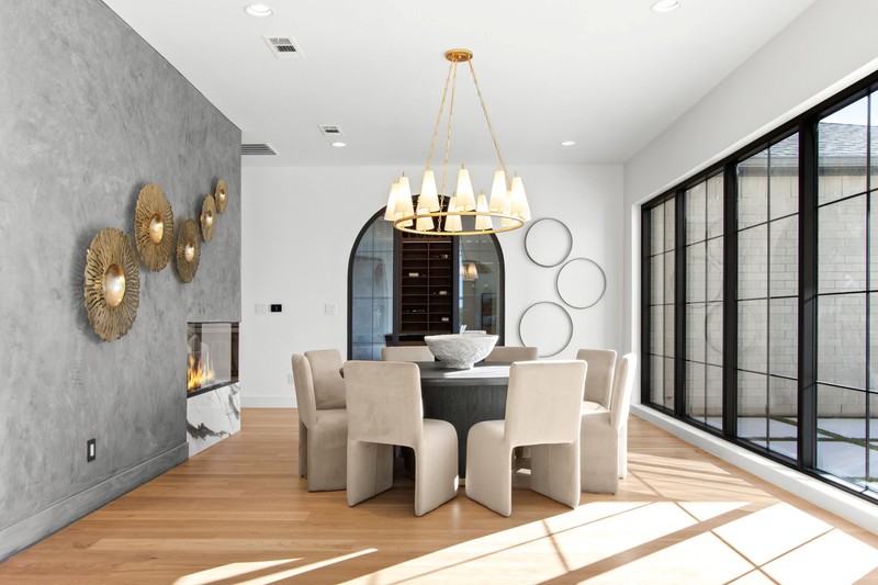 Modern dining area showcasing a round table with plush chairs, a stylish chandelier, and decorative wall elements. Natural light floods through large windows.