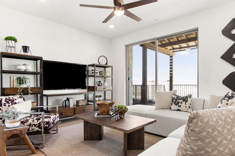 Modern living room featuring a white sectional, wooden coffee table, and bookshelf, with large windows opening to a balcony view.