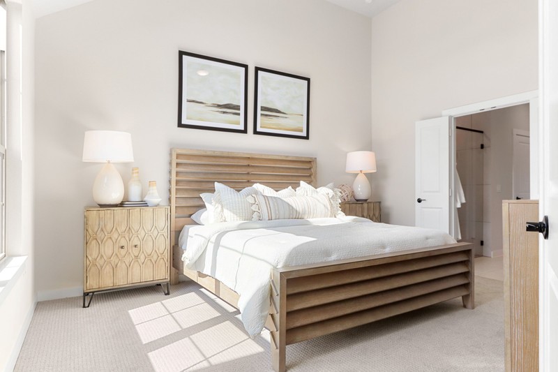Bright, airy bedroom featuring a wooden slatted bed, neutral bedding, and soft furnishings. Sunlight streams through large windows, enhancing a serene atmosphere.