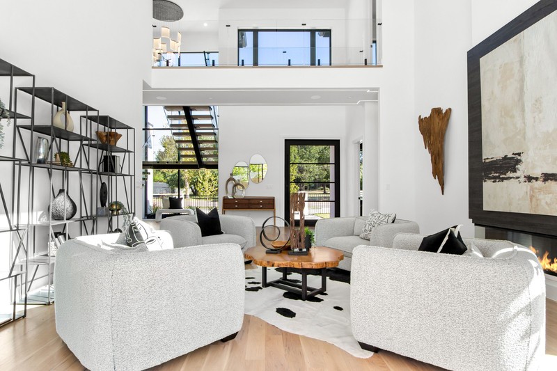 Modern living room with a high ceiling, featuring cozy white sofas, a wooden coffee table, and an artistic wall décor. Large windows showcase greenery outdoors.