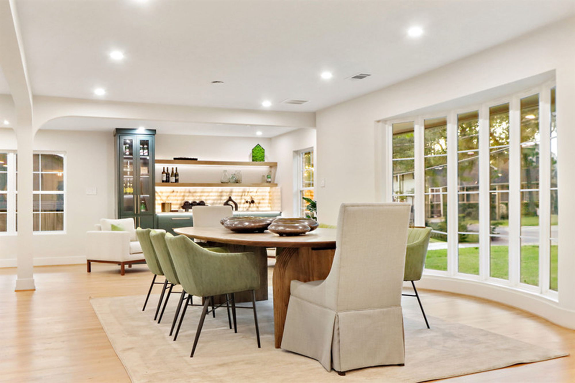 Bright, modern dining area featuring a round wooden table surrounded by green and beige chairs. Large windows provide ample natural light.
