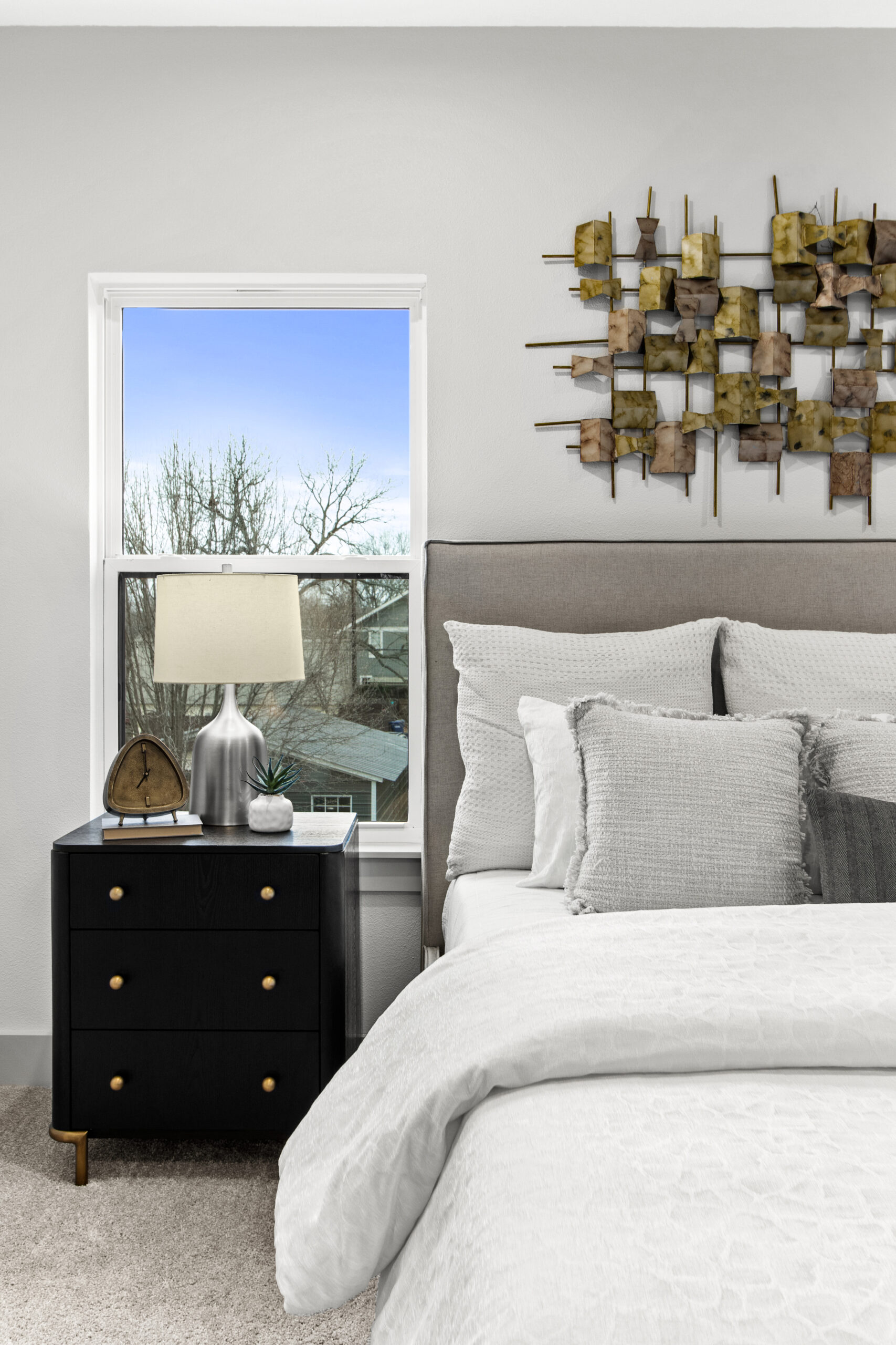Modern bedroom featuring a cozy bed with white bedding, a sleek black nightstand with a lamp, clock, and plant, and a large window showing trees outside.